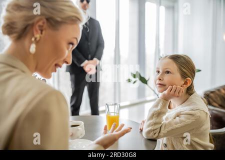 preteen kid listening to blonde mother attentively, discussing something in cafe, bodyguard on blurred background, security service, private safety co Stock Photo