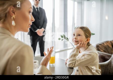 preteen girl listening to blonde mother attentively, discussing something in cafe, bodyguard on blurred background, security service, private safety c Stock Photo