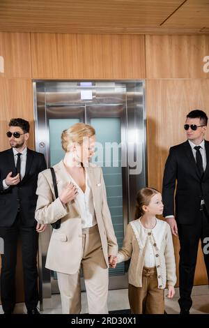 mother and child holding hands while standing near hotel elevators and bodyguards in suits, personal protection, successful woman and preteen daughter Stock Photo