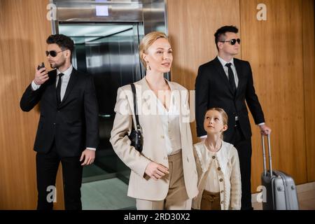 successful mother and child holding hands while standing near hotel elevator and bodyguards in suits, personal protection, woman and preteen daughter, Stock Photo