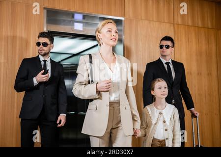 blonde mother and child holding hands while standing near hotel elevator and bodyguards in suits, personal protection, successful woman and preteen da Stock Photo