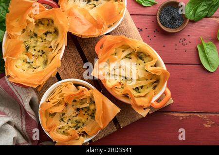 Filo pies with soft feta cheese and spinach in ceramic molds on old red wooden table backgrounds. Filo portions pies. Small Baked Spanakopita pies. To Stock Photo