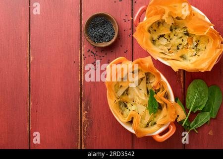 Filo pies with soft feta cheese and spinach in ceramic molds on old red wooden table backgrounds. Filo portions pies. Small Baked Spanakopita pies. To Stock Photo