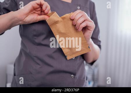 Close-up of women hands with a neat manicure opening a paper bag. A cosmetologist in a gray uniform tears open a bag of cosmetic tools. Doctor opens a Stock Photo