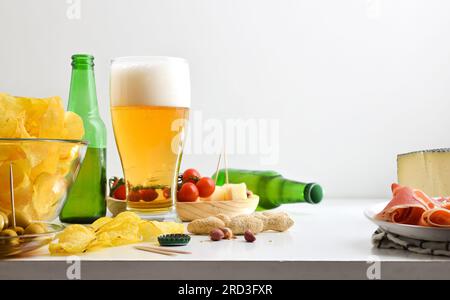 Appetizer with glass of beer and chips, peanuts, ham and cheese on white kitchen bench and white isolated background. Front view. Stock Photo