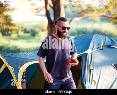 A brutal man with a beard in black glasses holds a plate with vegetables, cucumbers and tomatoes in his hand. Picnic in nature. Stock Photo