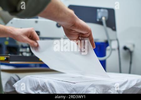 Cropped view of young craftsman putting layer on t-shirt and working on screen printing machine at background in print studio, customer-focused small Stock Photo