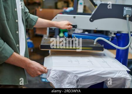 Cropped view of craftsman holding layer on t-shirt and working with screen printing machine in blurred workshop, customer-focused small business conce Stock Photo