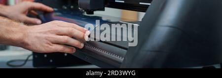 Cropped view of young craftsman holding layer while working with screen printing machine in blurred workshop, small business owner working on project, Stock Photo