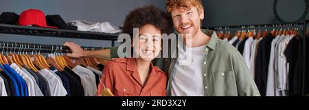 Smiling young multiethnic craftspeople looking at camera while standing together near clothes on hangers in print studio, young small business owners Stock Photo