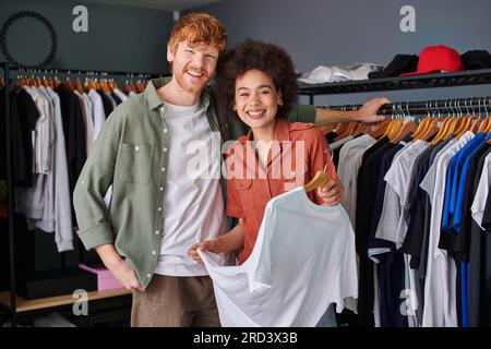 Positive young multiethnic craftspeople looking at camera while standing near clothes on hangers and working in print studio, young small business own Stock Photo