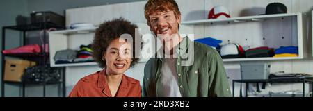Positive and young multiethnic craftspeople looking at camera while standing together and working in blurred print studio, ambitious young entrepreneu Stock Photo