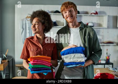 Smiling young multiethnic craftspeople holding clothes while standing and working together in blurred print studio at background, ambitious young entr Stock Photo