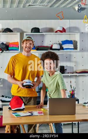 Cheerful young multiethnic designers looking at camera while working with snapbacks, laptop and cloth samples on table in print studio at background, Stock Photo
