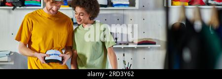 Cheerful young multiethnic craftspeople holding snapback with printing layer while working in blurred print studio at background, sustainable business Stock Photo