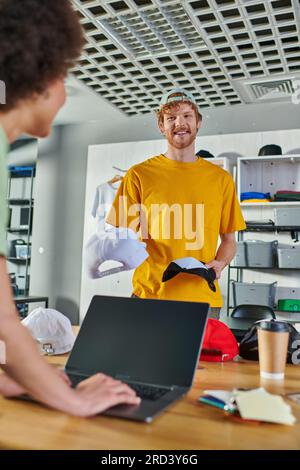 Cheerful young craftsman holding snapbacks and talking to blurred african american colleague using laptop and working on project in print studio, smal Stock Photo