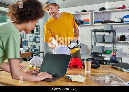 Smiling young designer holding snapbacks while talking to african american colleague using laptop near color swatches in print studio, small business Stock Photo