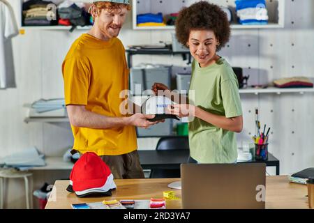 Smiling young multiethnic designers working with printing layer and snapback while looking at laptop near cloth samples in print studio at background, Stock Photo