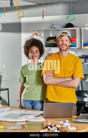 Smiling young multiethnic designers looking at camera and posing while standing near laptop and clothes during project in blurred print studio at back Stock Photo