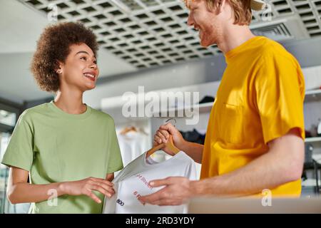 Smiling young multiethnic designers talking and looking at each other while holding t-shirt with lettering on hanger and working in print studio, star Stock Photo