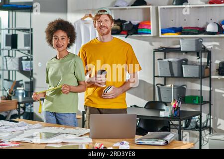 Positive multiethnic craftspeople holding coffee to go and tape measure while looking at camera and standing near laptop and clothes on table in print Stock Photo