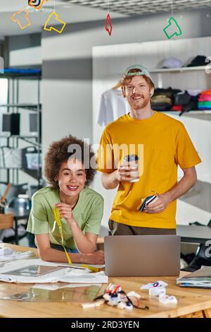 Smiling young multiethnic designers holding tape measure and coffee to go while looking at camera near laptop and clothes on table in print studio at Stock Photo
