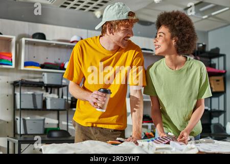 Cheerful multiethnic young designers talking while holding coffee to go and color swatches and working near clothes in blurred print studio at backgro Stock Photo