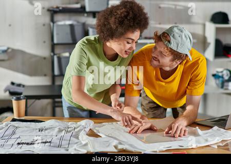 Positive young african american designer pointing with finger while working with colleague, printing layers and clothes near laptop in print studio, t Stock Photo