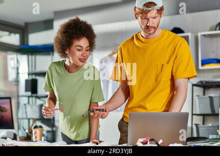 Smiling young african american craftswoman holding printing layer while working with colleague using laptop and clothes in print studio at background, Stock Photo