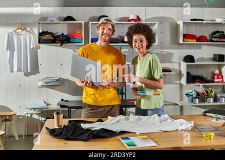 Positive young multiethnic craftspeople looking at camera while holding cloth swatches and laptop near clothes on table in print studio at background, Stock Photo