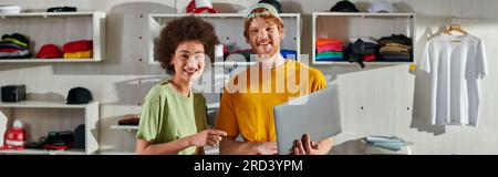 Positive multiethnic craftspeople looking at camera while working with laptop and standing near blurred clothes in print studio at background, small b Stock Photo