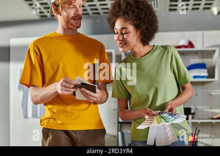 Smiling young artisan holding color swatches and talking to african american colleague while working together in blurred print studio at background, s Stock Photo