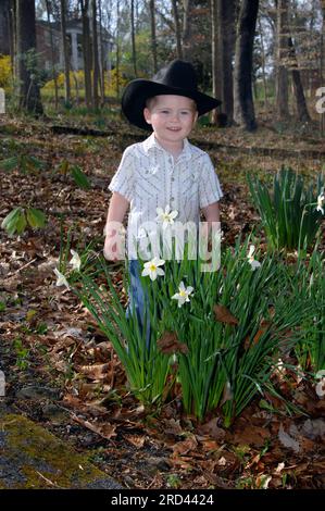 Black hat, jeans and snap up shirt makes this little boy feel like a cowboy.  He is standing besides blooming daffodils and is smiling. Stock Photo