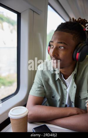 Young Man Commuting To Work Sitting  On Train Wearing Wireless Headphones Looking Out Of Window Stock Photo