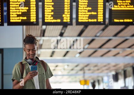 Young Man Man Wearing Backpack Looking At Mobile Phone By Train Departure Board In Railway Station Stock Photo