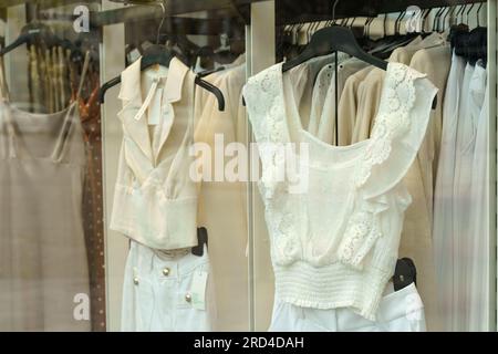 Light-colored women's clothing hangs on display racks behind glass in a shop window. Stock Photo