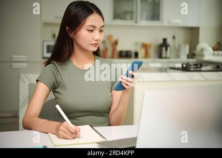 Serious college student checking notifications on smartphone when taking notes in textbook Stock Photo