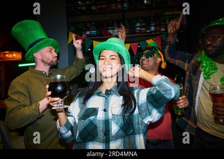 Young cheerful woman in grey flannel and green hat dancing among friends and looking at camera while enjoying St Patrick day party Stock Photo