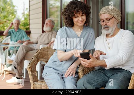 Young female caregiver pointing at screen of smartphone held by bearded senior man while explaining him how to use new mobile application Stock Photo