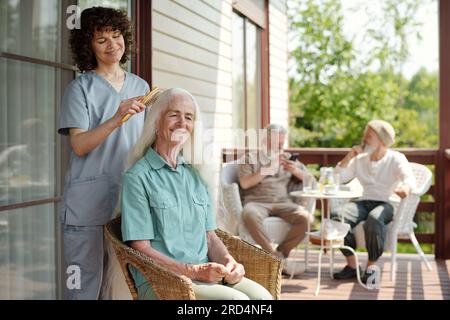 Happy young caregiver brushing long white hair of smiling female patient of retirement home on patio on summer morning against two aged men Stock Photo