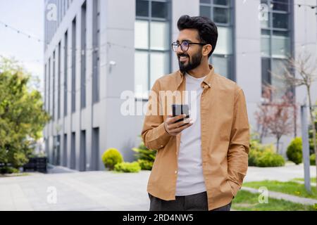 Successful young Hindu man walking outside office building, engineer software developer programmer smiling and happy using test app on phone, happy satisfied with result. Stock Photo