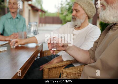 Focus on hand of senior man holding transparent cup with pills while sitting by wooden table in front of camera against other elderly people Stock Photo