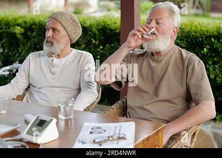 Bearded senior man taking pills in the morning while sitting by table next to another grey haired pensioner against green bushes in the garden Stock Photo