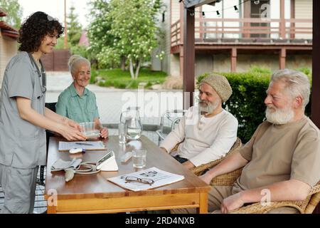 Happy young nurse bringing medicaments to group of senior people sitting by table on terrace of retirement home on summer morning Stock Photo