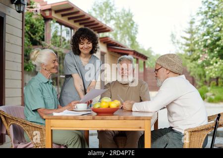 Young smiling carer in uniform and senior female patient of retirement home looking at paper document next to elderly bearded men Stock Photo