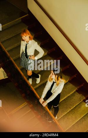 Two schoolgirls, dressed in their school uniforms, standing near the staircase railing during a school day. They are friends. Stock Photo