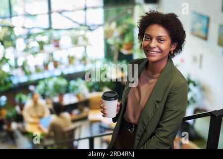 Young smiling African American businesswoman with cup of coffee looking at camera while standing against handrail of staircase in coworking space Stock Photo