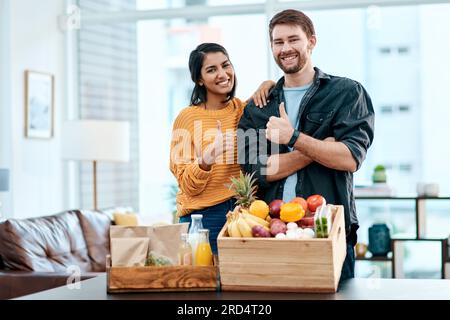Good health is the way to go. Shot of a happy young couple showing thumbs up after unpacking their healthy groceries at home. Stock Photo