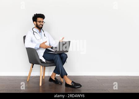 Handsome middle eastern young doctor sitting in armchair, using computer Stock Photo