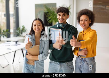 Happy diverse students gesturing thumbs up, standing in coworking space, approving their college educational program Stock Photo